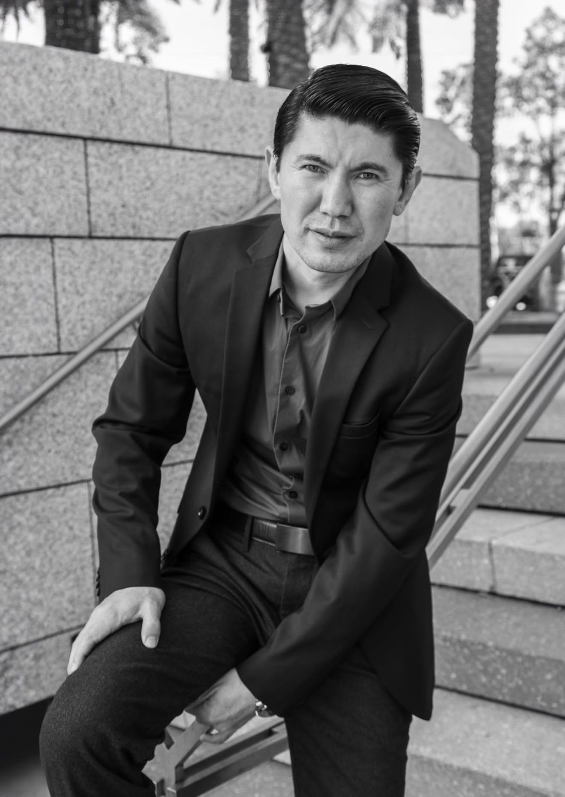 Black-and-white portrait of Medet Aman in a suit, seated on outdoor stairs