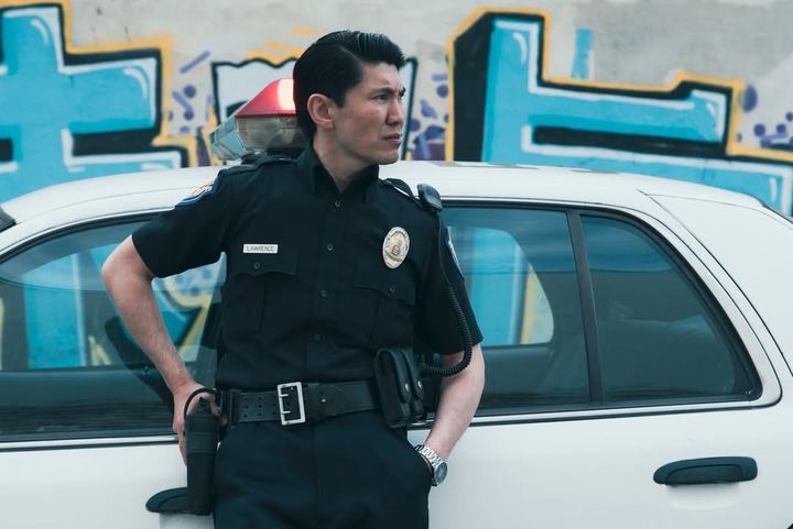 Medet in police uniform leaning on a patrol car, wide shot